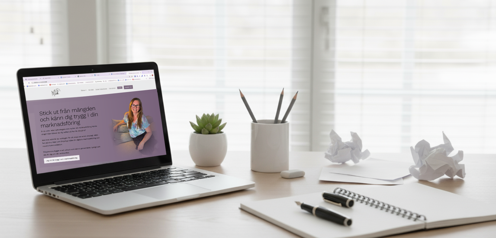 Ett skrivbord med en dator som visar startsidan på Mwh Kommunikation. Bredvid ligger ett block med en penna. 