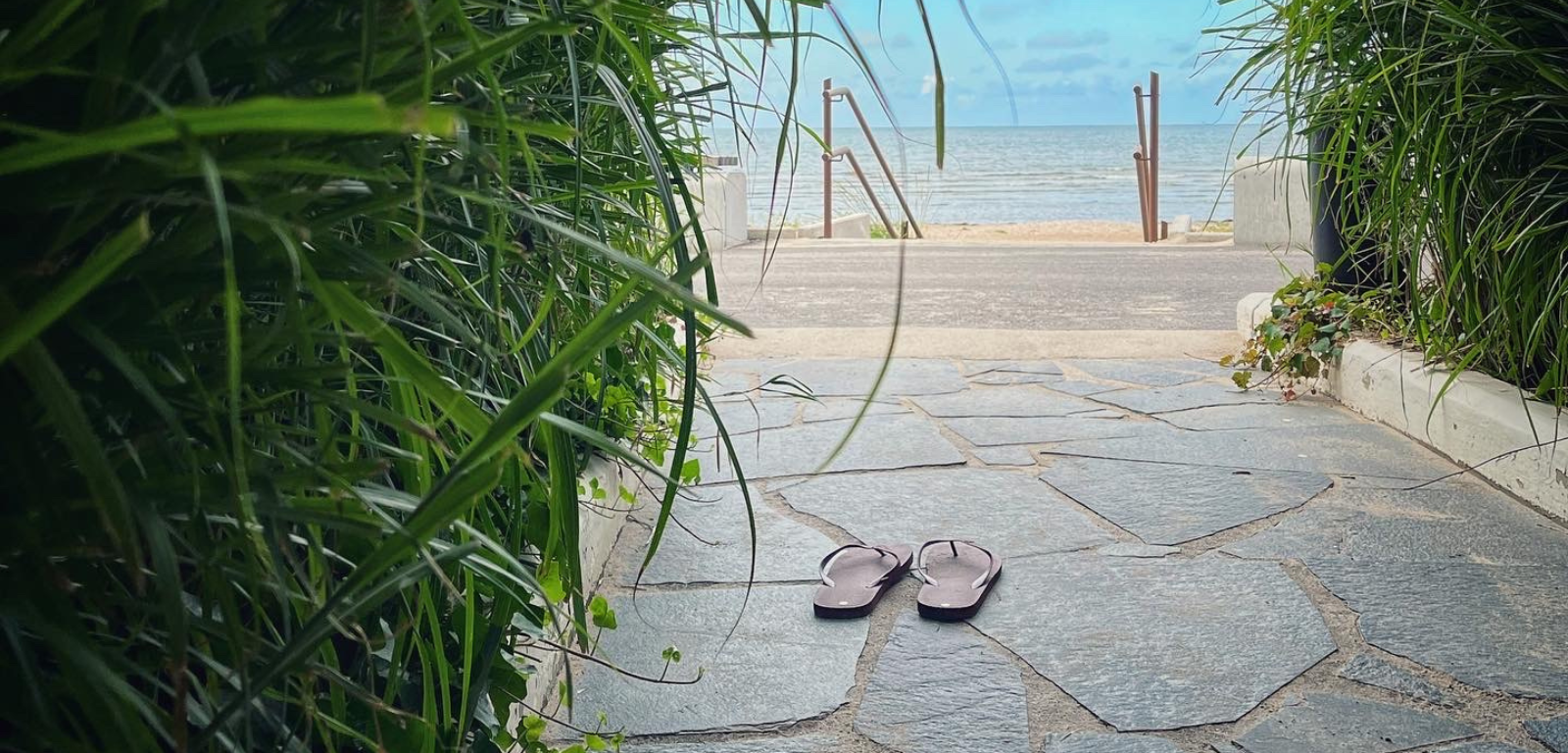 En gång med vass vid sidorna och ett par flip-flop som står på en gång. Längre bort syns havet och en sandstrand.